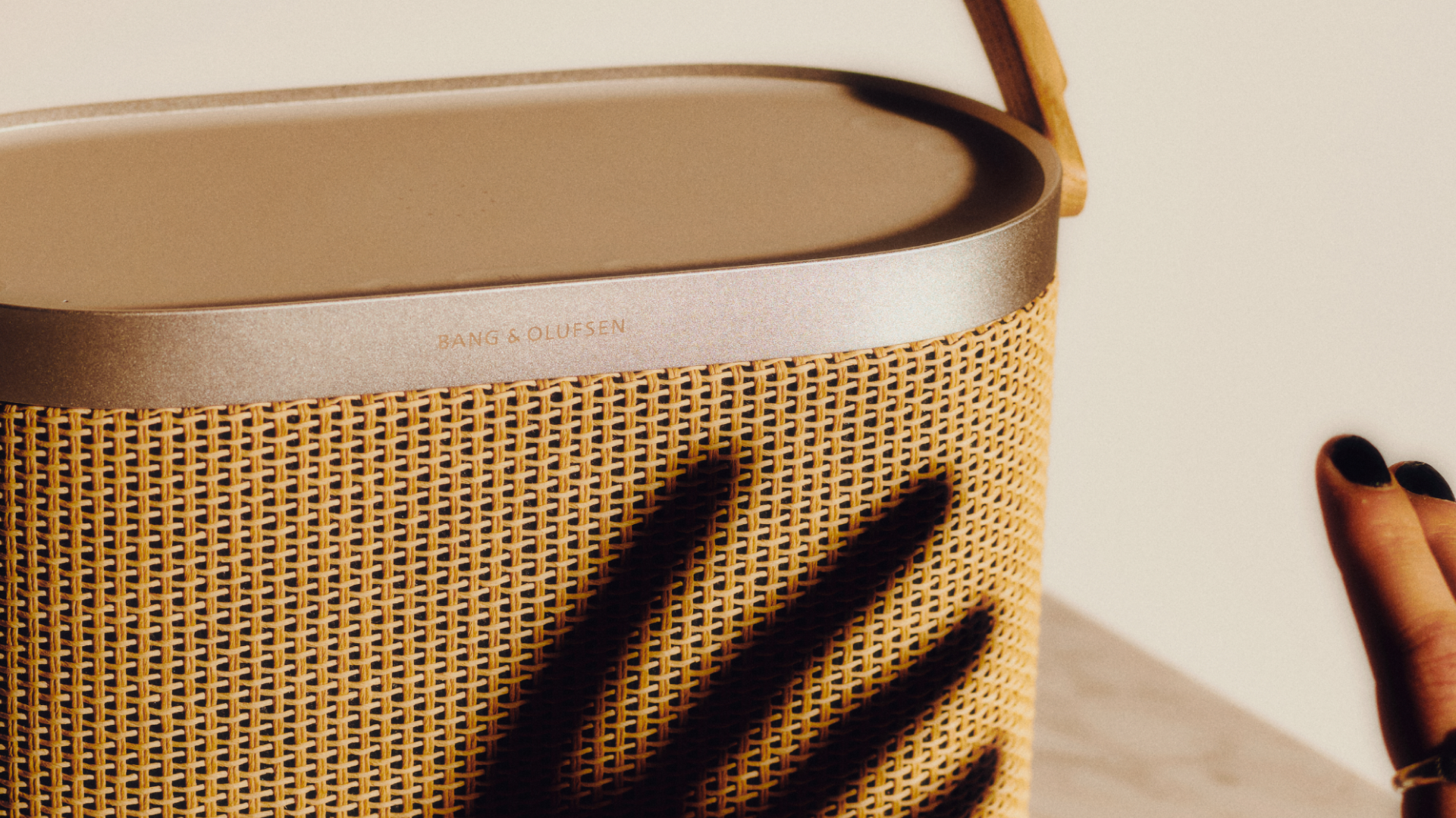 Nahaufnahme der Beosound A5 in der Nordic Weave Edition. Zu sehen ist das handgewebte Papiergeflecht der Front, der Rahmen aus natürlichem Aluminium mit eingraviertem Bang & Olufsen Logo und der Schattenwurf einer Hand.