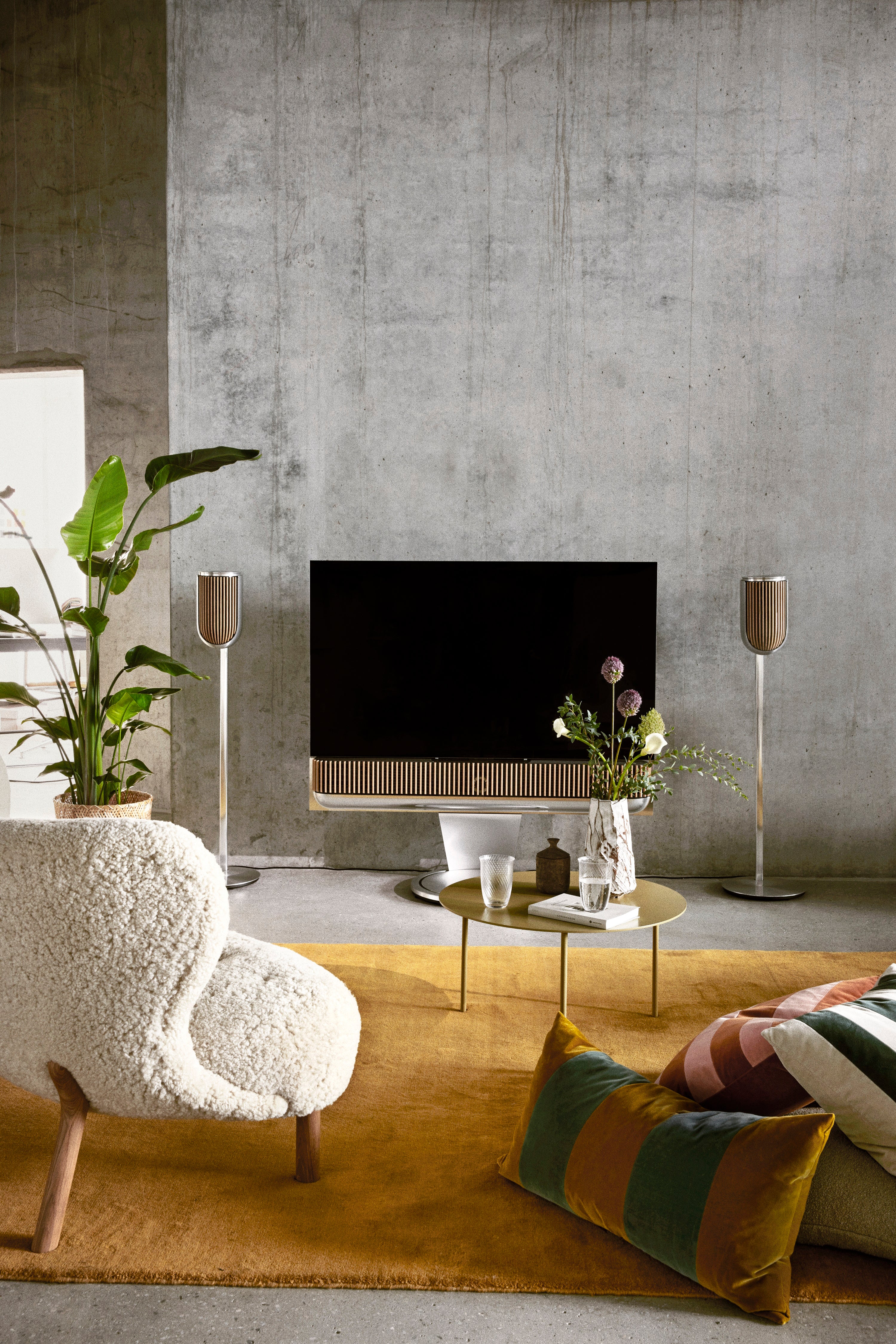 Moderne Wohnzimmer-Szene mit einer Bang & Olufsen Beovision Theater auf einem motorisierten Standfuß, flankiert von zwei Beolab 8 Lautsprechern auf hohen Bodenstandfüßen. Das Setup in Natural Aluminium und Eiche steht vor einer Sichtbetonwand, kontrastiert durch einen warmen gelben Teppich und hochwertige Designermöbel.