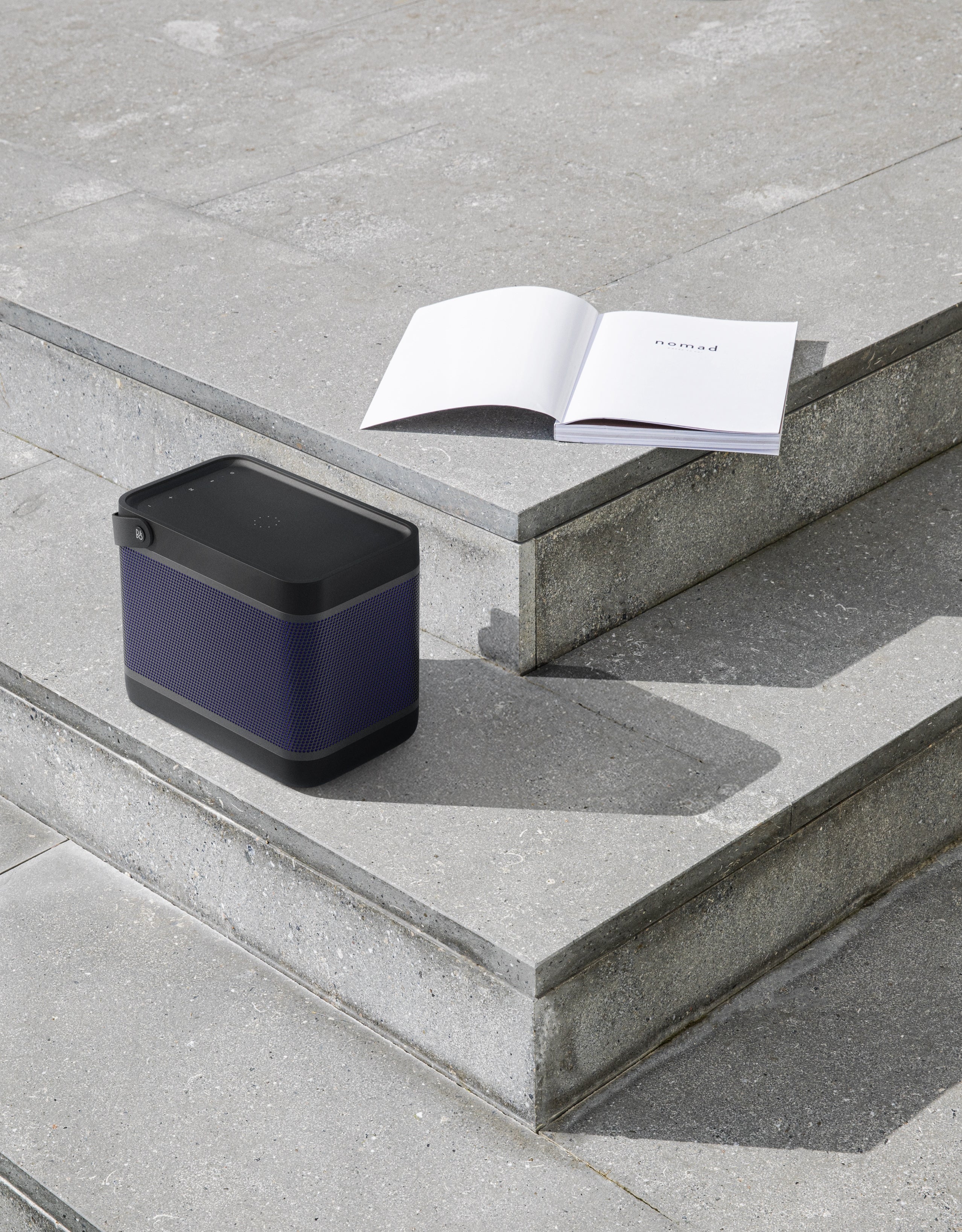 Minimalistische Lifestyle-Aufnahme des Bang & Olufsen Beolit 20 in black anthracite auf einer Betontreppe im Freien. Der portable Lautsprecher mit schwarzem Griff steht neben einem aufgeschlagenen Buch und unterstreicht das diskrete, skulpturale Design in einer modernen urbanen Umgebung.