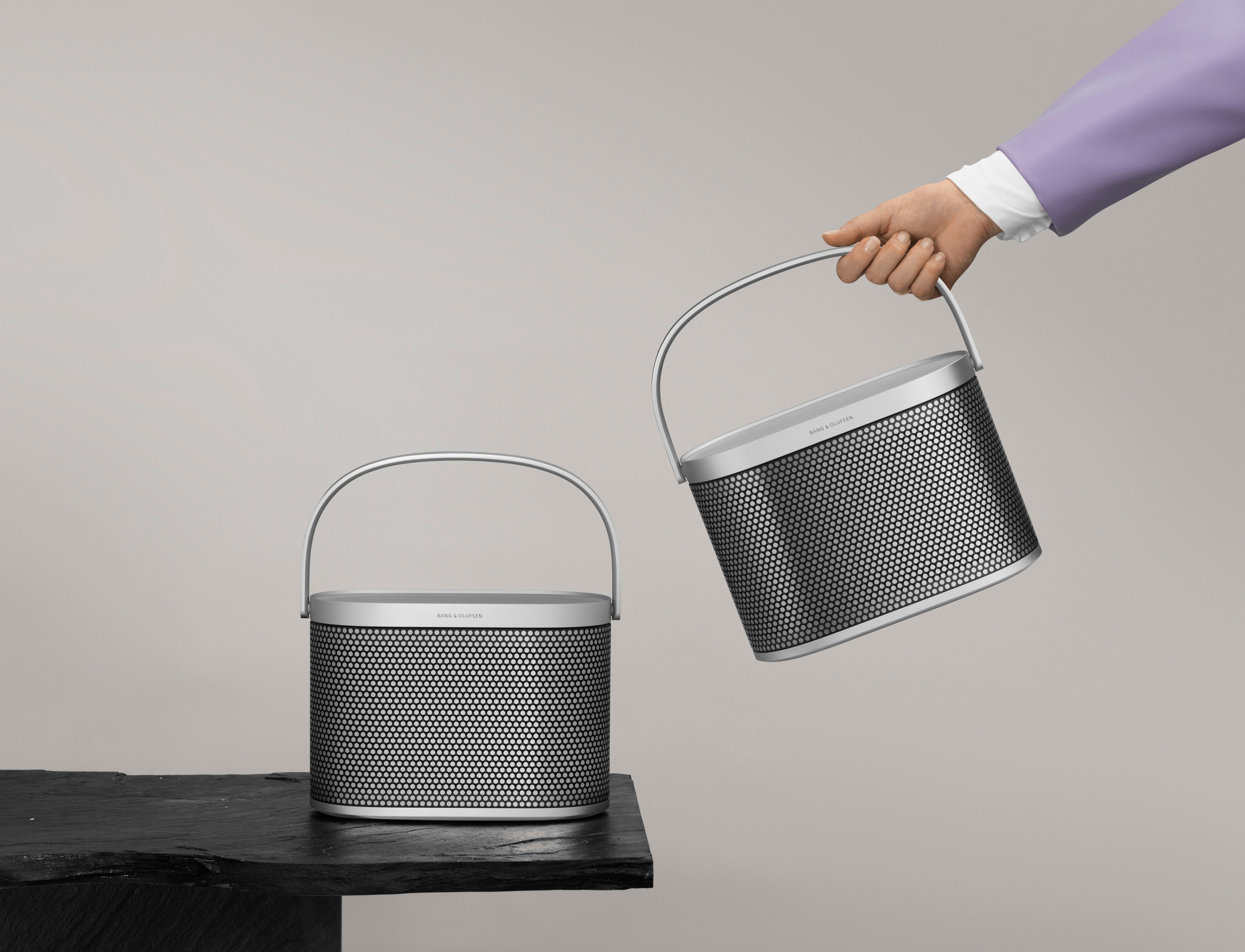 Zwei Beosound A5 Lautsprecher in der Spaced Aluminium Edition mit markantem Lochmuster. Ein Gerät steht auf einer dunklen Schieferplatte, das zweite wird von einer Hand am Aluminiumgriff angehoben, um das Stereo-Pairing und die Portabilität zu verdeutlichen.