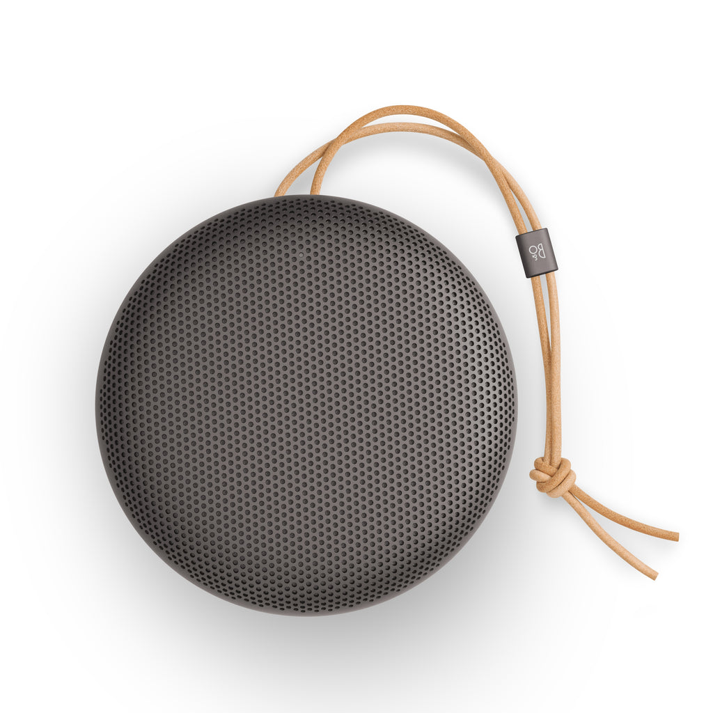 Draufsicht auf einen runden Bang & Olufsen Beosound A1 (3. Generation) Bluetooth-Lautsprecher in Warm Granite mit charakteristischem Aluminium-Gitter und einer hellbraunen Leder-Trageschlaufe vor weißem Hintergrund.