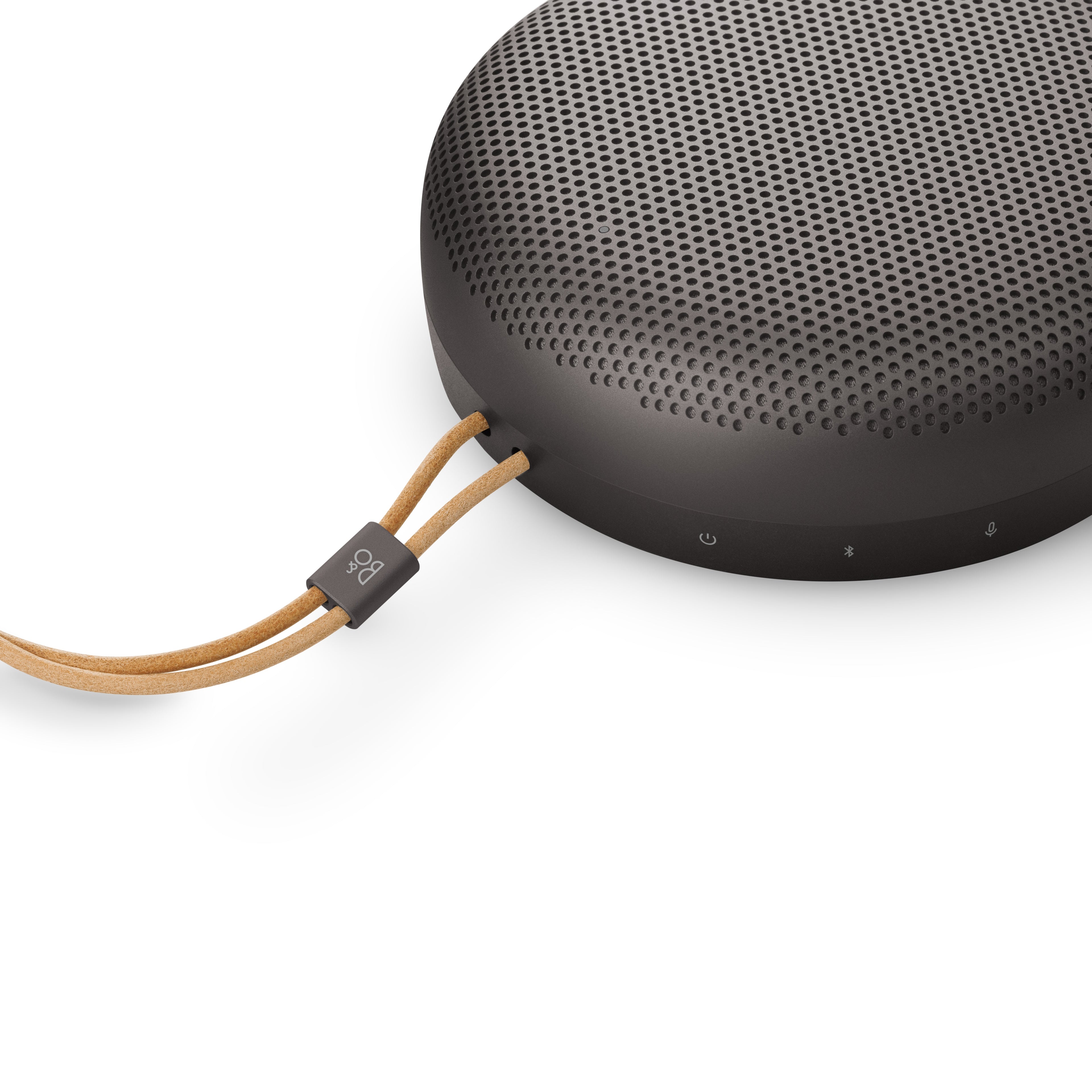 Detailansicht des Bang & Olufsen Beosound A1 von schräg oben, mit Fokus auf die hellbraune Lederkordel inklusive Logo-Tag sowie die Symbole für Power, Bluetooth-Pairing und Mikrofon-Stummschaltung am Gehäuserand.