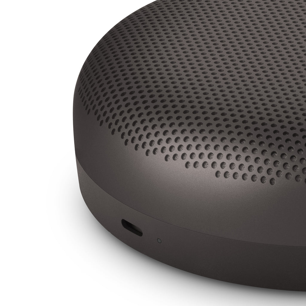 Nahaufnahme der Seitenansicht des Bang & Olufsen Beosound A1 in Anthrazit mit Fokus auf den USB-C Ladeanschluss, die LED-Betriebsanzeige und das präzise perforierte Aluminium-Gehäuse