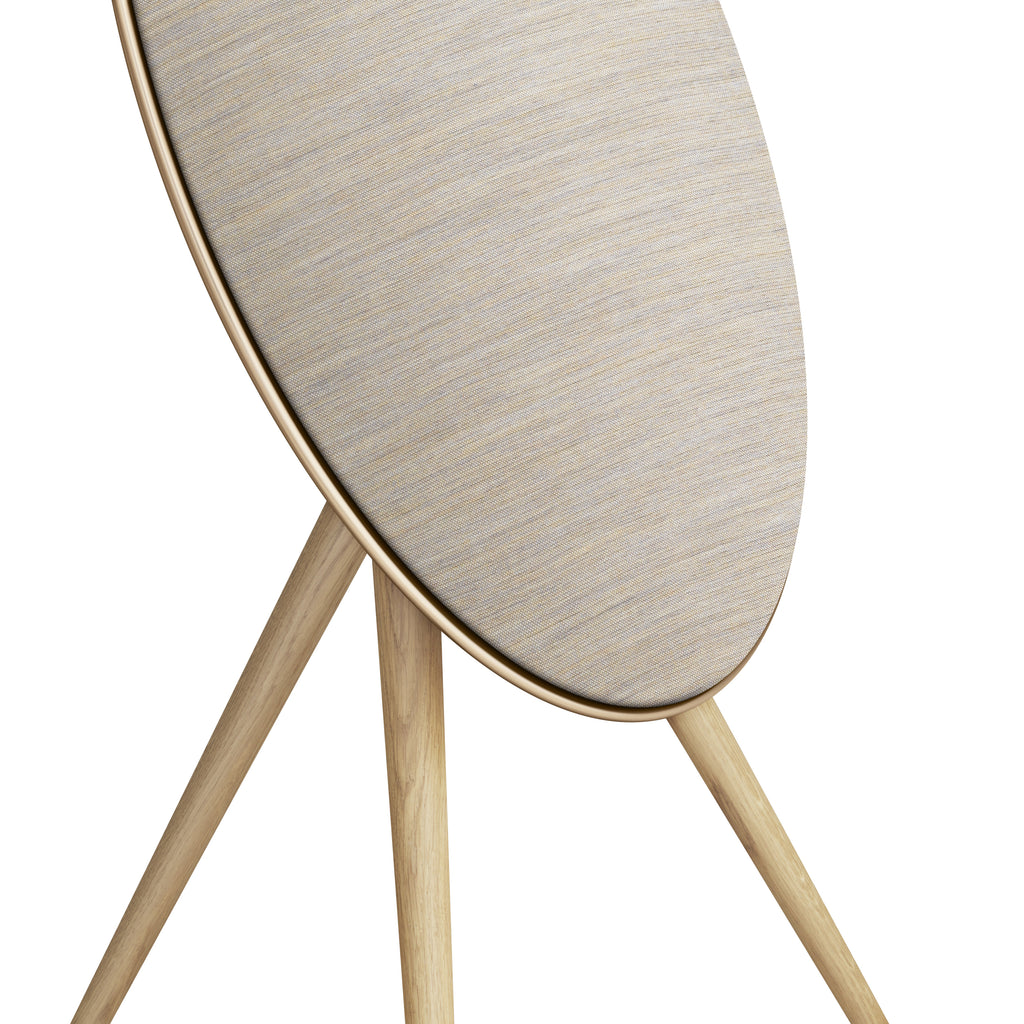 Detaillierte Nahaufnahme der Bang & Olufsen Beosound A9. Das Bild zeigt den Übergang zwischen dem goldfarbenen Aluminiumrahmen, der melierten Textilabdeckung von Kvadrat und den Beinen aus massiver heller Eiche. Ein Fokus auf die hochwertige Materialkombination und handwerkliche Präzision.