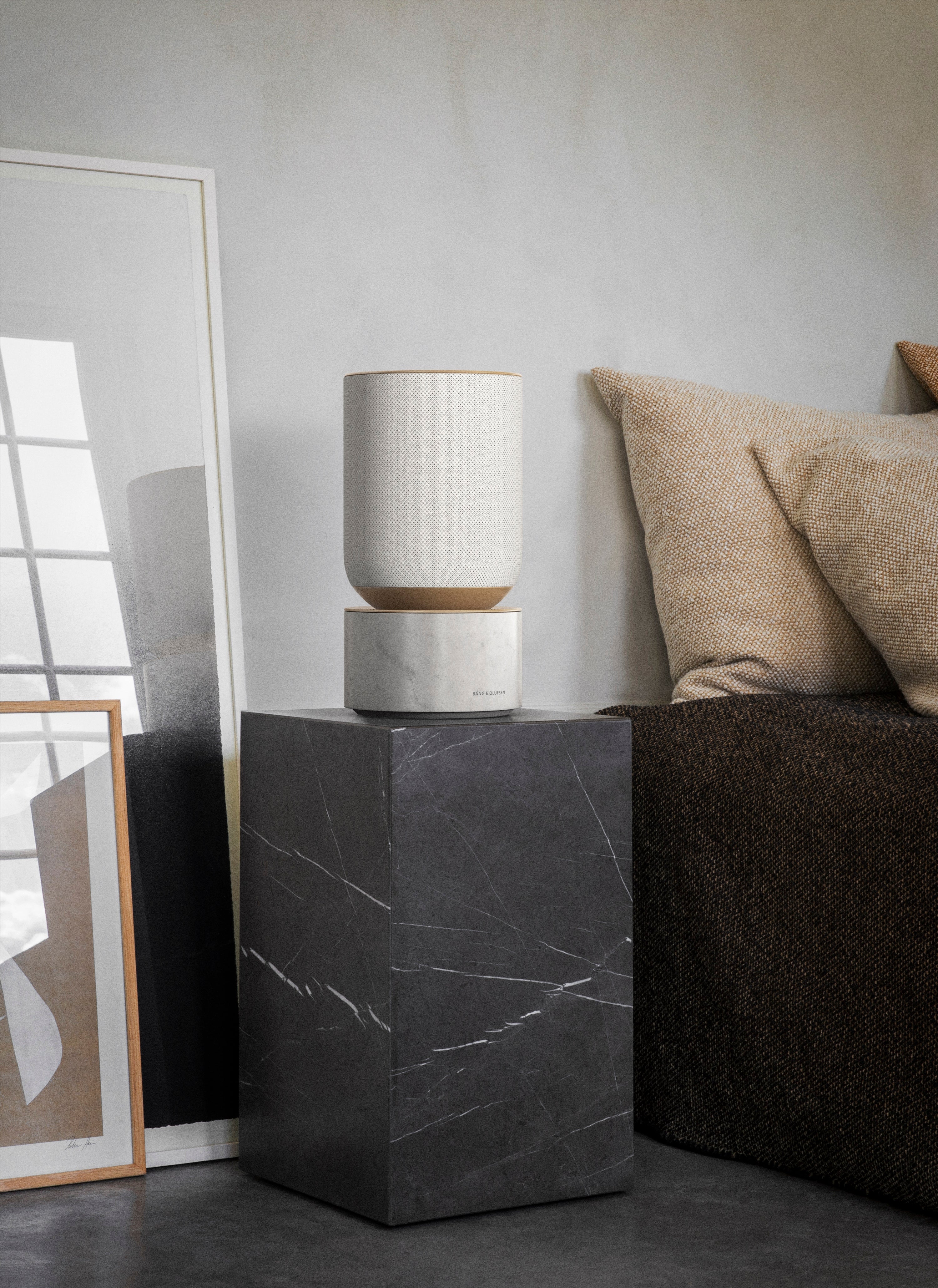 Lifestyle-Aufnahme des Bang & Olufsen Beosound Balance in der exklusiven White Marble Edition. Der Lautsprecher mit hellem Textil-Oberteil steht auf einem Sockel aus echtem Carrara-Marmor, platziert auf einer dunklen Marmorsäule in einem minimalistisch gestalteten Raum mit sanften Erdtönen.