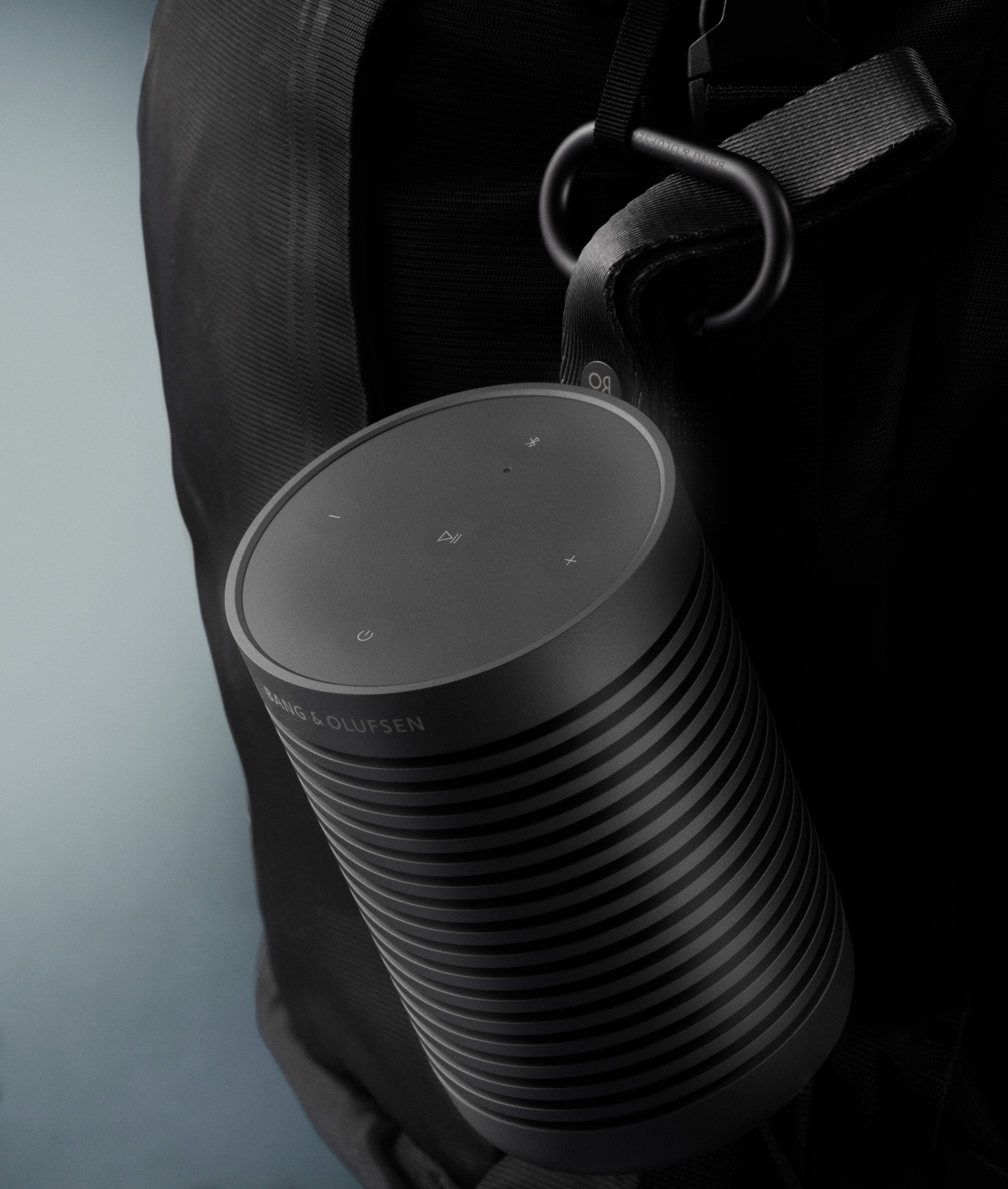 Lifestyle-Nahaufnahme des Beosound Explore in Black Anthracite. Der robuste, zylindrische Outdoor-Lautsprecher ist mit seinem schwarzen Riemen und einem Karabiner an einem taktischen Rucksack befestigt. Das Bild betont die Portabilität und das widerstandsfähige Design für den Einsatz unterwegs.