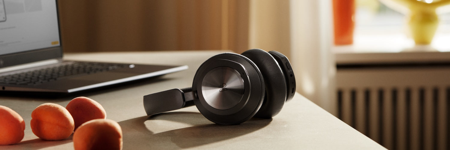 Lifestyle-Aufnahme der Bang & Olufsen Beocom Portal Over-Ear-Kopfhörer in Anthrazit auf einem hellen Schreibtisch. Im Hintergrund ist ein Laptop zu sehen, im Vordergrund frische Aprikosen. Das Bild betont das elegante Aluminium-Finish der Ohrmuscheln und die hochwertige Lederpolsterung in einer natürlichen Arbeitsumgebung.