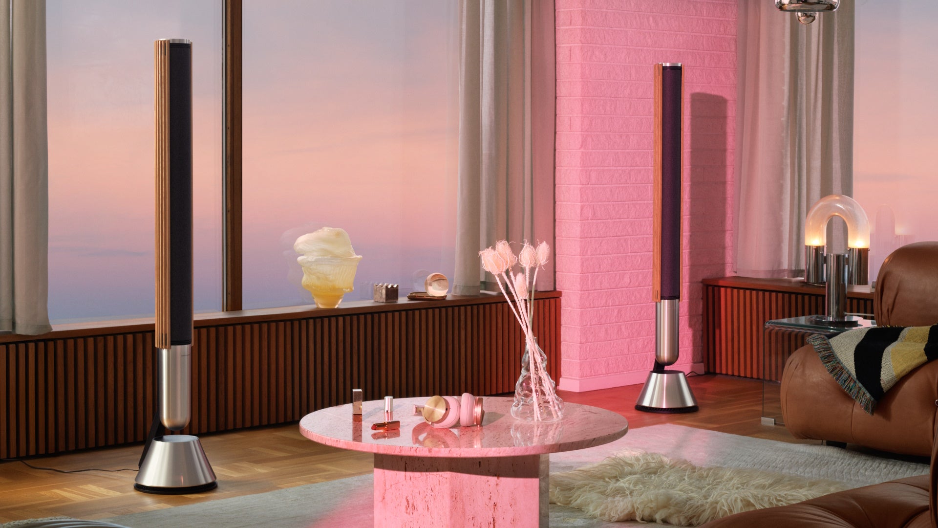 Panorama-Lifestyle-Aufnahme eines Stereo-Paars der Bang & Olufsen Beolab 28 Standlautsprecher in Silber mit hellen Eichenholz-Lamellen. Die Szene zeigt ein luxuriöses Wohnzimmer bei Sonnenuntergang, akzentuiert durch pinkfarbenes Licht, mit den Beoplay H95 Kopfhörern auf einem runden Marmortisch im Vordergrund.