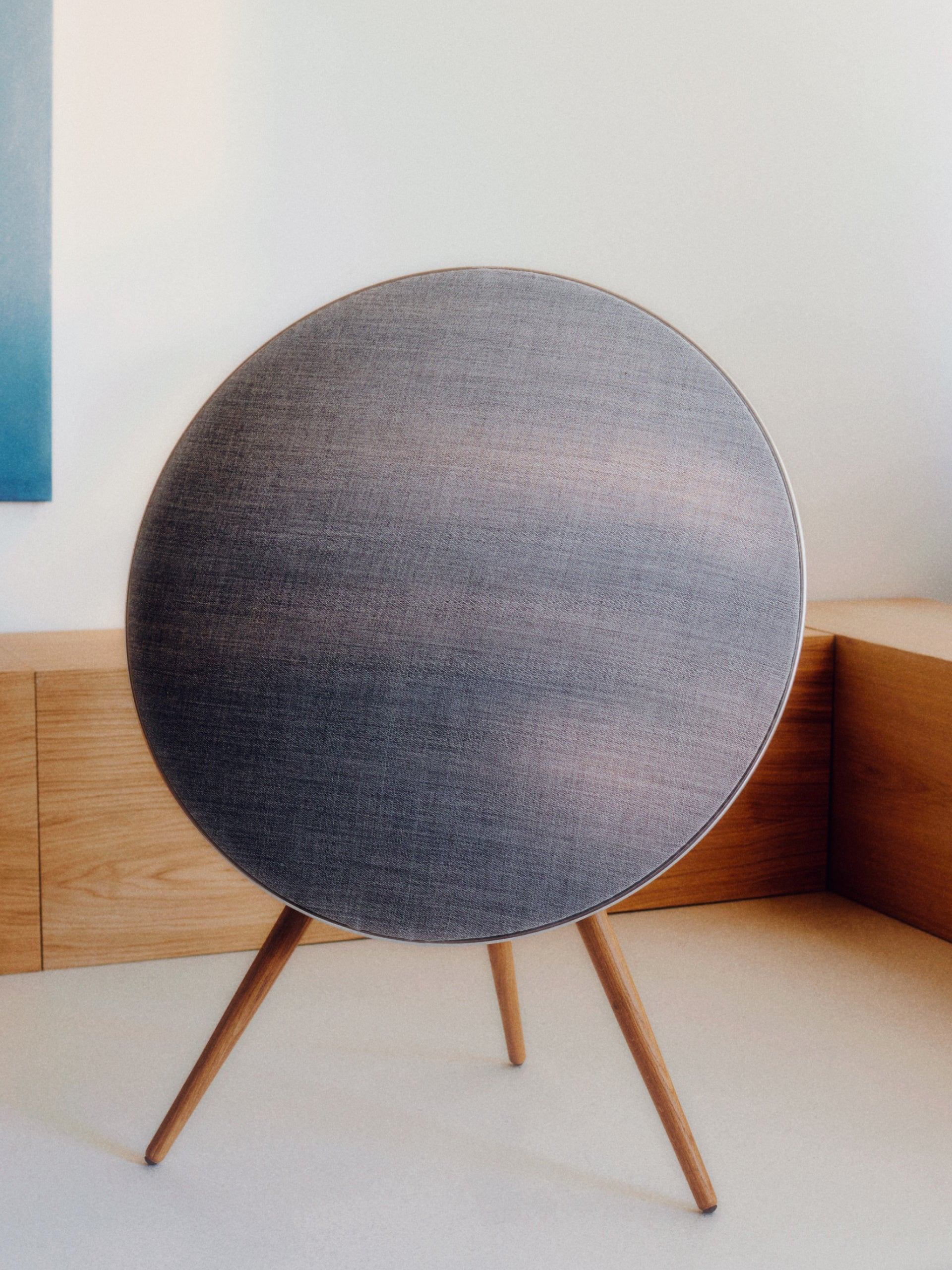 Lifestyle-Aufnahme der Bang & Olufsen Beosound A9 in einem minimalistischen Interieur. Der Lautsprecher mit silbernem Aluminiumrahmen, dunkelgrauem Melange-Stoffbezug und Eichenholzbeinen steht vor einer passenden Holz-Schrankwand. Das Bild vermittelt reale Proportionen und die harmonische Integration in modernes Wohndesign.