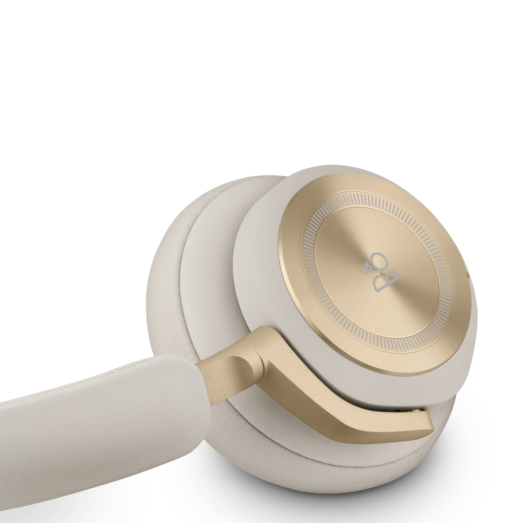 Detailaufnahme des Bang & Olufsen Beoplay HX in Sand. Fokus auf die goldfarbene Aluminium-Ohrmuschel mit feiner Rillenstruktur und dem B&O-Logo, die an einem sandfarbenen Lammleder-Polster befestigt ist.
